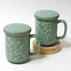 May include: Two matching green ceramic mugs with lids. Each mug has a handle and a floral pattern. The mugs are on a light-colored surface. The lids have the same floral pattern as the mugs.