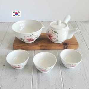 Peut inclure: Un service à thé en céramique blanche comprenant une théière avec une poignée latérale, un bol verseur et trois petites tasses à thé. Chaque pièce est ornée d'un motif floral peint à la main en rose et vert, disposé sur une planche en bois.