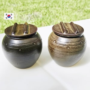 May include: Two ceramic jars with lids. The jars are dark brown and have a rounded shape with a lid on top. The lids have a handle. The jars are sitting on a white surface with green grass in the background.