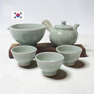 May include: A ceramic tea set featuring a light green, speckled glaze with a crackle finish. The set includes a side-handled teapot with a lid, a wide pouring bowl, and three small tea cups, all arranged on a rectangular wooden block.