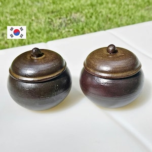 May include: Two small, round, ceramic jars with lids. The jars are dark brown and have a glossy finish. The lids are a lighter brown color and have a small knob on top. The jars are sitting on a white surface with green grass in the background.