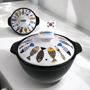 Puede incluir: Una olla de cerámica negra con una tapa blanca que presenta un patrón colorido de varios peces y el texto Blue Fish. Se muestra una tapa a juego en el fondo. El diseño incluye motivos de peces amarillos, azules y negros sobre fondo blanco.