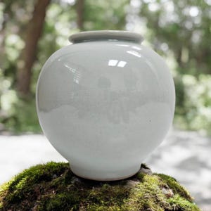 Pode incluir: Um vaso de cerâmica redondo e bulboso com um esmalte cinza-claro liso e brilhante. O recipiente apresenta um pescoço curto e estreito com uma borda circular plana e uma base ligeiramente cônica, exibindo uma silhueta limpa e uma superfície reflexiva.