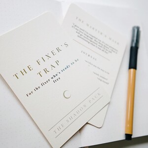 Puede incluir: Tarjetas blanquecinas con texto dorado que dice "THE FIXER'S TRAP" y "THE SHADOW PACK". Un bolígrafo negro y marrón descansa sobre un cuaderno blanco. Las tarjetas tienen texto legible. El tema es la escritura de diarios y la autorreflexión.