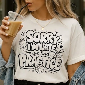 May include: White t-shirt with the text "SORRY I'M LATE we had PRACTICE" in a black, playful font. The design includes a basketball, a whistle, and an alarm clock. The person is wearing a denim jacket and holding a coffee drink.