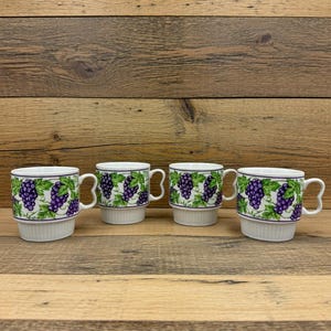 May include: Four white ceramic mugs with a grape design. The mugs have a white base with a ribbed texture and a handle. The upper portion of the mugs features a pattern of green leaves and purple grapes.