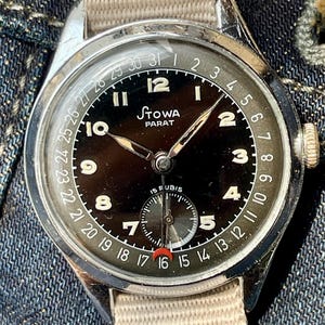 Może przedstawiać: Zegarek na rękę Stowa Parat w stylu vintage z czarną tarczą, świecącymi wskazówkami i sub-tarczą na godzinie 6. Zegarek posiada kopertę ze stali nierdzewnej i beżowy pasek z tkaniny. Tarcza wyświetla datę i tekst "STOWA PARAT".