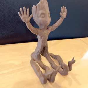 May include: A light brown figurine of a character with a tree-like appearance, arms raised, and a smiling face. The figurine has a textured surface and is posed with its arms up and its legs spread out.