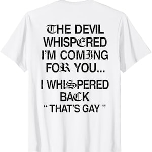 May include: White t-shirt with black text. The text reads "THE DEVIL WHISPERED I'M COMING FOR YOU... I WHISPERED BACK 'THAT'S GAY'" in a bold, gothic font. The shirt has short sleeves and a crew neck.