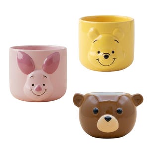 Könnte beinhalten: Drei Keramikbecher mit Winnie-Puuh-Figuren. Einer ist gelb mit Puuhs Gesicht, ein anderer ist rosa mit Ferkels Gesicht und der dritte ist braun mit einem Bärengesicht. Jeder Becher hat eine abgerundete Form.