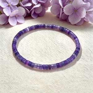 Pode incluir: Uma pulseira circular composta por pequenas contas planas em forma de disco em vários tons de roxo, variando do lavanda claro ao violeta profundo. As contas estão dispostas num círculo contínuo, criando uma banda fina e flexível.
