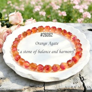 Könnte beinhalten: Eine Halskette aus facettierten, orangefarbenen Achatperlen, kreisförmig angeordnet, liegt auf einer weißen, strukturierten Oberfläche. Der Text "#26062", "Orange Agate" und "a stone of balance and harmony" ist sichtbar.