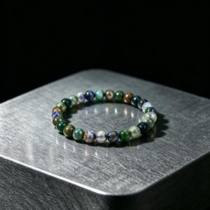 May include: A circular beaded bracelet composed of small, polished spherical stones in various shades of deep green, earthy brown, and mottled blue. The beads are strung together in a continuous loop, displaying natural colour variations and textures.