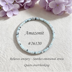 May include: A circular bracelet made of small, flat, disc-shaped beads in pale blue with black speckled inclusions. The text Amazonit #26120 is centered within the circle, followed by the text Relieves anxiety - Soothes emotional stress and Quiets overthinking.