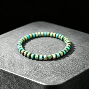 Puede incluir: Una pulsera circular compuesta por pequeñas cuentas planas en forma de disco en tonos variados de azul turquesa y marrón terroso. Las cuentas están ensartadas en un cordón fino de color turquesa a juego, creando una banda continua y flexible.