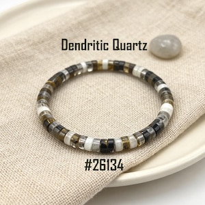 May include: A circular bracelet composed of small, flat, disc-shaped beads made of Dendritic Quartz. The beads feature a repeating pattern of translucent, opaque white, and dark brown to black tones. The text Dendritic Quartz and #26134 are visible.