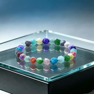 Op de afbeelding: Een kleurrijke kralenarmband met facetgeslepen edelstenen in verschillende kleuren, waaronder rood, groen, blauw en paars. De armband wordt gepresenteerd op een glazen oppervlak, dat het licht en de kleuren van de kralen weerspiegelt. Een sieraad.