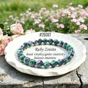 Pode incluir: Uma pulseira de contas de Ruby Zoisite facetadas, em tons de verde e roxo. A pulseira é exibida em um prato branco. O texto diz "Ruby Zoisite" e "boost vitality, ignite creativity, balance emotions."