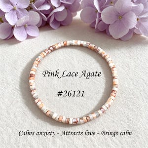 May include: A circular bracelet made of small, flat, disc-shaped pink lace agate beads with marbled white and peach tones. The text "Pink Lace Agate", "#26121", and "Calms anxiety - Attracts love - Brings calm" is printed in the center.