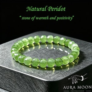 以下が含まれることがあります： 緑色のペリドットブレスレット、カットビーズ付き。「Natural Peridot」と「stone of warmth and positivity」の文字。ブレスレットは金属の表面に表示されています。