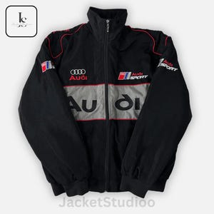 May include: Black zip-up jacket with red and gray accents. Features the Audi logo and "Audi Sport" branding. The jacket has a high collar and a full-length zipper. The text "JacketStudioo" is at the bottom.