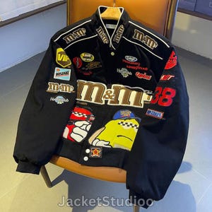 May include: Black bomber jacket with multiple patches and embroidered logos, including "M&M's" and "38". The jacket features a white collar and trim, with a button-up front. The jacket is displayed on a wooden chair.