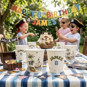 May include: A pirate-themed birthday party table setting featuring white paper cups printed with the name James and the number 8, a miniature wooden ship model, a small treasure chest filled with gold coins, and a Jolly Roger banner.