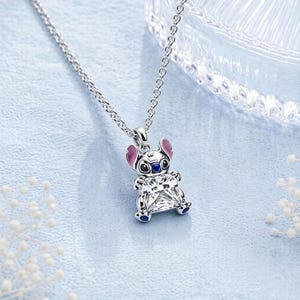 May include: A silver-toned metal necklace featuring a pendant shaped like the character Stitch. The pendant has pink enamel on the ears, blue enamel on the nose and feet, and holds a large, clear, square-cut faceted gemstone in its center.