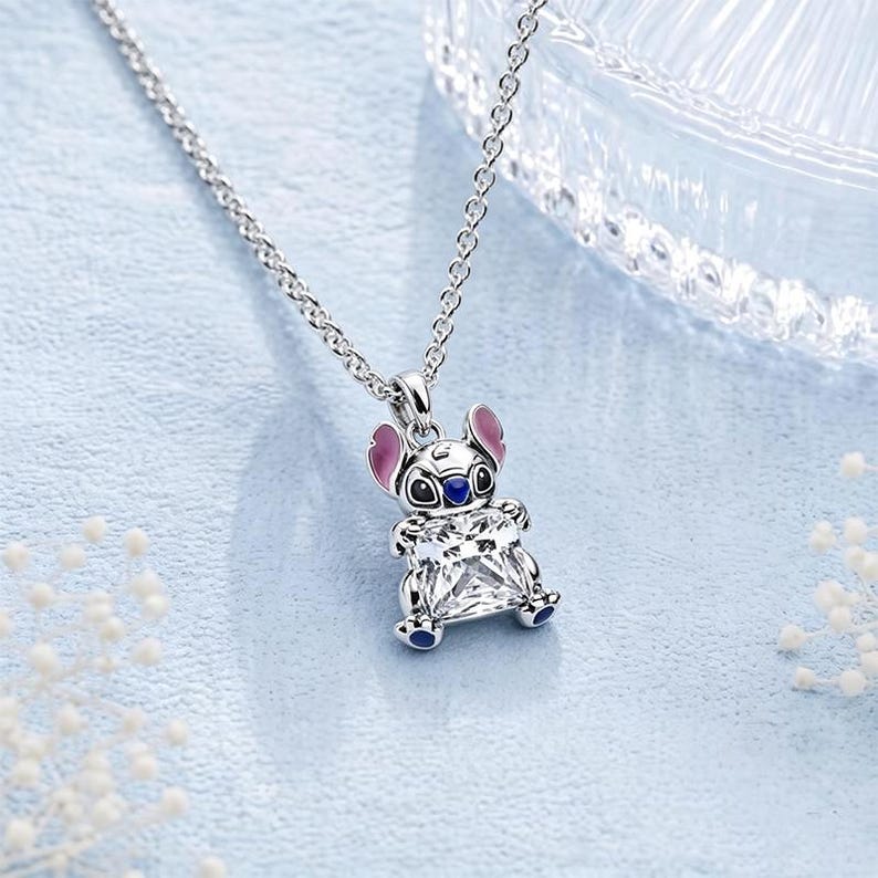 May include: A silver-toned metal necklace featuring a pendant shaped like the character Stitch. The pendant has pink enamel on the ears, blue enamel on the nose and feet, and holds a large, clear, square-cut faceted gemstone in its center.