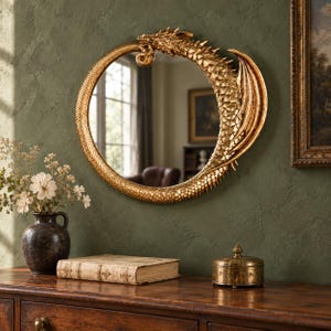 May include: A circular wall mirror framed by a sculpted gold dragon. The dragon's body forms the frame, featuring detailed scales, a head at the top, and a wing extending along the right side. The mirror surface reflects a room with a window.