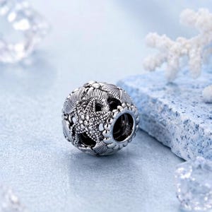 May include: A spherical silver charm bead featuring a detailed embossed starfish design surrounded by textured seashell patterns. The bead has a polished metallic finish with darkened recessed areas to highlight the intricate marine-themed carvings.