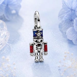 May include: A silver nutcracker charm featuring a blue enamel hat with a central star and a clear stone, red glitter enamel sleeves, and black enamel details on the face and torso, suspended from a polished silver jump ring bail.