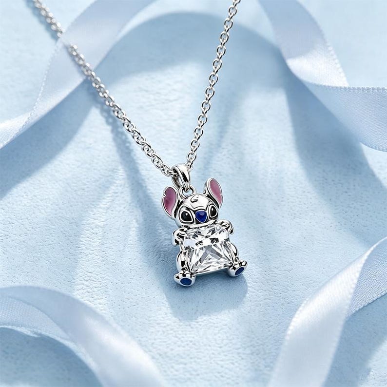 May include: A silver-toned pendant necklace featuring a character with large ears, pink inner ear accents, and dark blue nose and paw details. The character holds a large, square-cut clear gemstone in its center, suspended from a delicate chain.