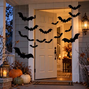 May include: A collection of black bat-shaped cutouts suspended by thin strings in front of an open doorway. The bats vary in size and wing position, creating a flying effect. The scene includes pumpkins and a lantern on the porch floor.