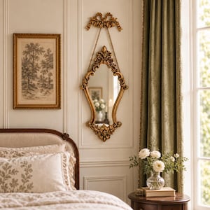 May include: An ornate gold wall mirror with a decorative bow top and hanging chain. The mirror features intricate scrollwork and a pointed frame design. It is mounted on a white paneled wall above a bed with patterned bedding and a bedside table.