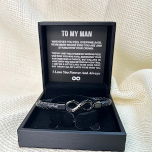 May include: A black leather bracelet with an infinity symbol charm, displayed in a black gift box. The infinity charm has the text "Carlos & Kelly" engraved on it. The box includes a message that reads "To My Man".