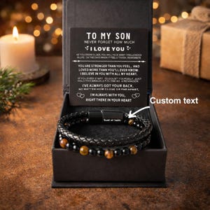 Puede incluir: Una pulsera de cuero negro con cierre magnético, presentada en una caja de regalo negra. La pulsera presenta una banda de cuero trenzado y una hebra de cuentas negras y marrones. Una tarjeta dentro de la caja dice "TO MY SON, I LOVE YOU."