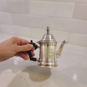 May include: A small, silver-colored teapot with a black handle and spout. The teapot has a decorative lid and a classic design. The handle is black and angular. The teapot is likely made of metal and is designed for serving tea.