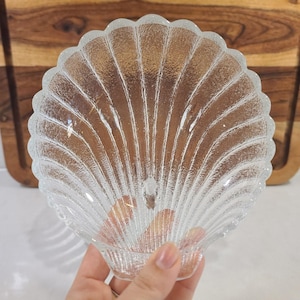 May include: Clear glass seashell-shaped dish with textured surface and scalloped edges. The dish is held in a hand, showcasing its delicate design. The dish is likely used for serving or display.