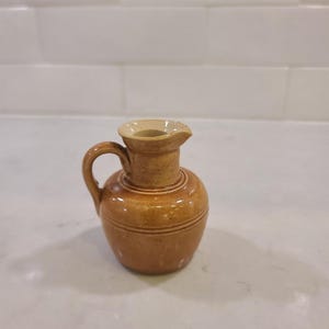 May include: A small, brown ceramic pitcher with a handle and a spout. The pitcher has a rounded body with horizontal lines and a lighter-colored rim. The pitcher is likely used for pouring liquids.