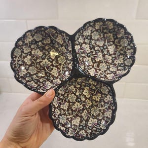 Puede incluir: Un plato para servir de tres secciones con un estampado floral. El plato tiene un fondo marrón oscuro con diseños florales blancos, morados y azul claro. Los bordes son festoneados y delineados en negro.