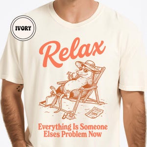 May include: Ivory-colored t-shirt with the word "Relax" in coral lettering. The shirt features a cartoon opossum in sunglasses, a hat, and a drink, lounging in a beach chair. The text "Everything Is Someone Elses Problem Now" is below the opossum.