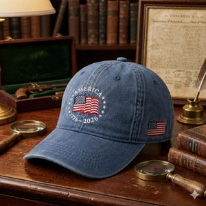 May include: A blue denim baseball cap with the word "AMERICA" embroidered in white, along with a US flag design and the years "1776-2026". A smaller US flag is embroidered on the side. The cap is on a wooden surface.