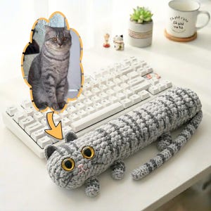 May include: A handmade grey and white striped crochet cat-shaped pillow with large yellow eyes and a small pink nose. The pillow is on a white keyboard. An image of a cat is in the background.