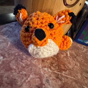 May include: A crocheted fox head in bright orange, with black and white details. The fox features black eyes and a white muzzle. The ears have black tips and white inner markings. The craftwork is visible, showcasing the textured yarn.