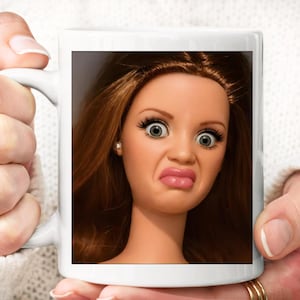 Może przedstawiać: Biały ceramiczny kubek z nadrukiem twarzy lalki z długimi brązowymi włosami, szerokimi szarymi oczami i wydętymi ustami w kolorze różowym. Lalka ma mały srebrny kolczyk w jednym uchu oraz gładką, jasną karnację.