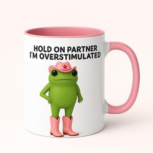 Può includere: Tazza in ceramica bianca con interno e manico rosa. La tazza presenta una rana verde dei cartoni animati con cappello e stivali da cowboy rosa. Il testo sulla tazza recita "HOLD ON PARTNER I'M OVERSTIMULATED."