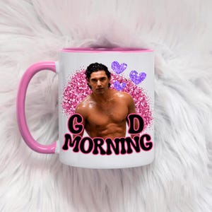 Op de afbeelding: Een witte keramische mok met een roze handvat en rand. De voorzijde toont een fotoprint van een persoon met ontbloot bovenlijf, omringd door een roze glitterende cirkelvormige achtergrond, drie paarse glitterende hartjes en de zwarte tekst G D MORNING.