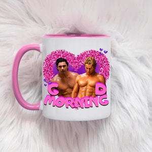May include: A white ceramic mug with a pink handle and rim featuring a graphic of two shirtless men inside a large pink glittery heart. The text "GOOD MORNING" is printed in bold pink letters below the heart, with small purple hearts above.