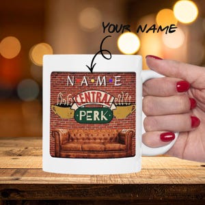 Può includere: Tazza in ceramica bianca con il logo "Central Perk" della serie TV Friends, con sfondo di muro di mattoni e divano in pelle marrone. La tazza ha un manico e la scritta "YOUR NAME" sopra il logo.